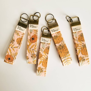 May include: Five keychains with floral patterns and gold-colored key rings. Each keychain has a pink background with orange flowers and green leaves. The text "fern & arrow" is printed on each keychain.