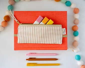 May include: A bright orange rectangular book with a beige and white striped fabric pencil case attached to the front. Three colorful markers peek out from the zipper. Below the book are three pens in pink, brown, and yellow. A felt ball garland surrounds.