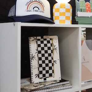 May include: A white and black checkered notebook with a palm tree design. The notebook is on a white shelf with other notebooks and a black and white checkered pouch.