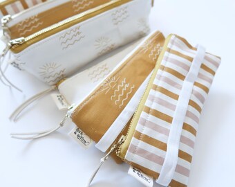 May include: Set of three zippered pouches with a white background and a tan stripe pattern. The pouches have a sun and wave design and the text "forth & willow" printed on the bottom.