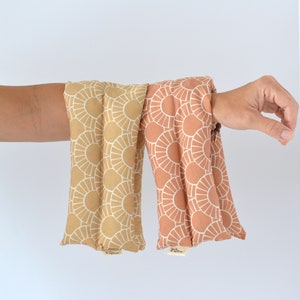 May include: Two fabric-covered heat packs with a geometric sunburst pattern. One is beige with white sunbursts, the other is peach with white sunbursts.