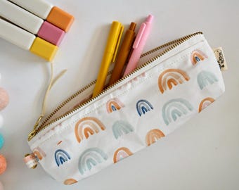 May include: A white canvas pencil case with a gold zipper and a repeating pattern of small rainbows in muted orange, blue, and green tones. The case contains three pens in yellow, brown, and pink, and features a small tag labeled Sun & Arrow.