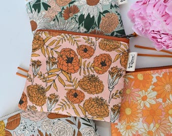 May include: A square fabric zipper pouch featuring a light pink background with a repeating pattern of orange marigold flowers and green leaves. The pouch has a rust-colored zipper and a small white fabric tag with the text fern & arrow.