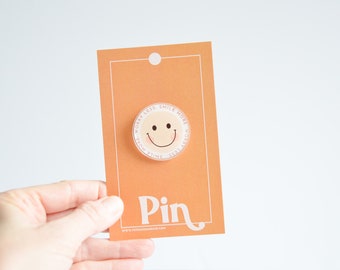 May include: A round, peach-colored pin with a white smiley face and the text "Worry Less Smile More" in a pink circle around the smiley face. The pin is on a peach-colored card with the word "Pin" in white.