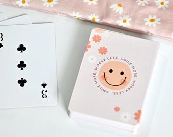 May include: A deck of playing cards featuring a light pink back design with a central peach-colored smiley face, small white and orange flowers, and the text "Worry Less. Smile More." A single three of clubs playing card is visible next to the deck.
