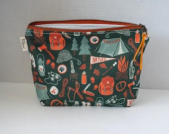 May include: A dark green zippered pouch featuring a repeating pattern of orange and white camping gear, including tents, backpacks, lanterns, axes, and a pennant with the word NATURE. The pouch has a rust-colored zipper and a yellow leather pull.