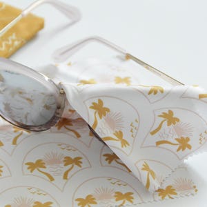 May include: Sunglasses with a light pink frame and gold arms rest on a white cloth. The cloth has a repeating pattern of gold palm trees, suns, and waves. A yellow square cloth is in the background.