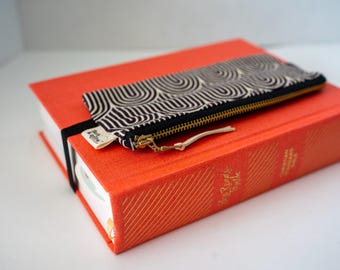 May include: A rectangular fabric pouch with a black and white arched pattern, a gold-toned metal zipper, and a leather pull tab, secured by a black elastic band to a bright orange hardcover book with gold embossed text on the spine.