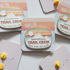 May include: Three enamel pins with a white background and a pastel pink, orange, and blue design. The pins feature the text "Pacific Northwest Trail Crew Chase Views Not Dudes".