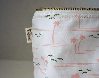 May include: A white fabric pouch featuring a repeating pattern of light pink palm trees and small dark green birds in flight. The pouch has a gold-toned metal zipper at the top and a small white fabric tag on the side with the text fern & arrow.