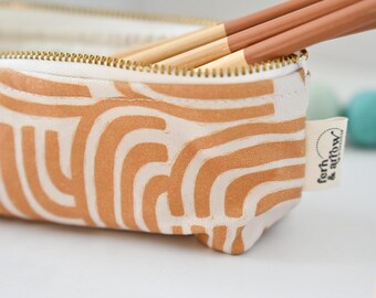 May include: A rectangular fabric pencil case featuring a cream background with a tan abstract line pattern. The case has a gold-toned metal zipper and a small white fabric tag on the side with the text ferh & arrow printed in black ink.