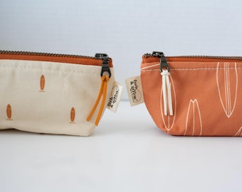 May include: Two rectangular zippered pouches featuring a surfboard pattern. The left pouch is cream with orange surfboards and an orange zipper. The right pouch is orange with white surfboard outlines and a white zipper pull. Both have a brand tag.
