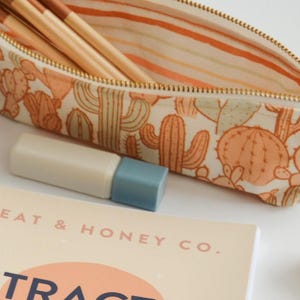 May include: A white zippered pencil case with a cactus print. The case is open and has a gold zipper. The inside of the case is lined with orange and white stripes. The case is filled with wooden pencils.