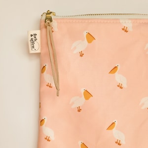 May include: A peach-colored zippered pouch featuring a repeating pattern of white pelicans with orange beaks and feet. A small white fabric tag on the side displays the text fern & arrow workshop. The zipper pull includes a thin tan leather cord.