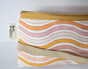 May include: A rectangular fabric pouch featuring a repeating pattern of wavy lines in shades of mustard yellow, soft pink, and cream. The pouch has a mustard yellow zipper at the top and a beige elastic band across the front, with a white tag reading fern & arrow.