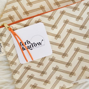 May include: A white zippered pouch with a brown geometric pattern. The pouch has an orange zipper and a small orange tag with the text "fern & arrow workshop" on it.
