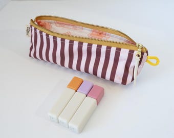 May include: A rectangular pencil case with vertical dark brown and light pink stripes, a gold zipper, and a yellow pull tab. The interior lining features a light-colored pattern. Three rectangular lip balms with white bases and orange, lavender, and pink caps.