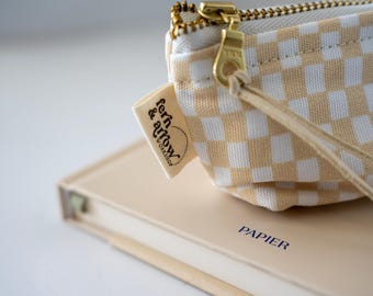 May include: A small beige and white checkered canvas pouch with a gold-toned metal zipper and a long leather pull. A fabric tag attached to the side displays the text fern & arrow WORKSHOP. The pouch rests on a beige book with the word PAPIER printed.