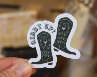 May include: A die-cut sticker featuring two dark green cowboy boots with white line art details of stars, moons, and leafy vines. The text GIDDY UP! is printed in a curved, dark green font above the boots, all set against a white background.
