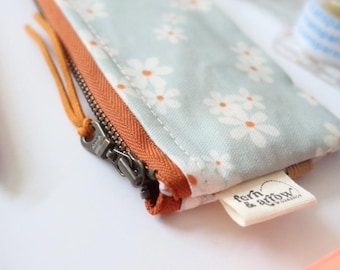 May include: A light blue floral print pencil case with a rust-colored zipper and pull. The case features a daisy pattern and a small tag that reads "fern & arrow". A pink pen is visible in the background.