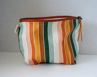 May include: A zippered pouch featuring a vertical striped pattern in shades of orange, mustard yellow, forest green, cream, and soft pink. The pouch has a rust-colored zipper closure with a thin leather pull and a small white fabric tag on the side.