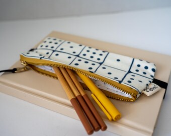 May include: A rectangular zippered pencil case featuring a white fabric with a dark blue domino pattern. The case has a mustard yellow zipper and contains three pens, including two brown pens and one mustard yellow pen, resting on a beige notebook.
