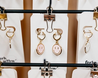 May include: A display of gold hoop earrings with various charms, including pink flowers, smiley faces, and chain links. The earrings are displayed on white cards with clips.