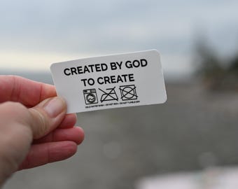May include: A white rectangular tag with rounded corners, held by a hand. The tag features the text "CREATED BY GOD TO CREATE" in black, along with laundry symbols. The bottom of the tag reads "COLD WATER WASH - DO NOT IRON - DO NOT TUMBLE DRY."
