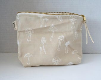 May include: A beige zippered pouch featuring a repeating white line-art pattern of various mushrooms. The bag has a gold-toned metal zipper closure with a matching fabric pull tab and a small white rectangular tag on the side with black text.
