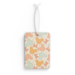May include: Rectangular air freshener with a repeating pattern of Mickey Mouse heads and bows in pastel colors. The background is a cream color with small star accents. A white string is attached for hanging.