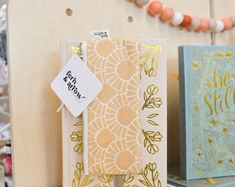 May include: A cream-colored book with gold floral foil stamping features a peach and white sunburst patterned fabric pencil case attached with an elastic band. A white tag with the text fern & arrow is attached to the zipper pull.
