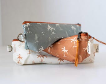 May include: Three rectangular fabric pencil cases stacked together. The cases feature a repeating pattern of white palm trees and surfboards on sage green, terracotta orange, and cream backgrounds. Each case has a zipper closure and a metal D-ring.