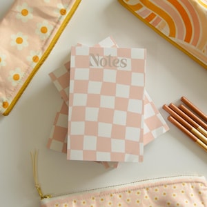 May include: A collection of stationery items, including notebooks, pencil cases, and pencils. The notebooks have a pink and white checkered pattern with the word "Notes". The pencil cases feature daisy and rainbow designs. The pencils are brown and gold.