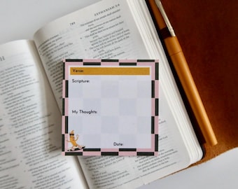 May include: A square, lined note card with a pink, black, and gold checkered border, resting on an open book. The card has spaces for "Verse", "Scripture", "My Thoughts", and "Date". A pen lies nearby.