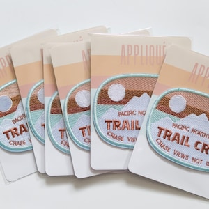 May include: Five iron-on patches with a light blue background and a brown, orange, and white mountain scene. The patches have the text "Pacific Northwest Trail Crew Chase Views Not Dudes" in a copper-colored font.