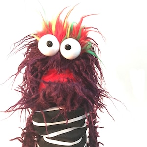 May include: Purple fuzzy puppet with red mouth and large white eyes. The puppet has a colorful mohawk and is wearing a black and white striped shirt.