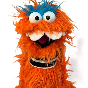 Könnte beinhalten: Orangefarbener und blauer Fell-Puppen mit großen Augen und einem weit geöffneten Mund. Die Puppe trägt ein schwarz-weiß gestreiftes Halsband.