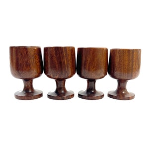 May include: Four dark brown wooden goblets. Each goblet features a rounded bowl, a slender stem, and a circular base. The wood grain is visible, adding texture. The goblets are arranged in a row against a white backdrop. A set of vintage drinkware.