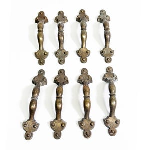 Op de afbeelding: Acht antieke messing lade handgrepen met een decoratief ontwerp. Elke handgreep heeft een centrale handgreep met een gedetailleerde boven- en onderplaat. Het messing heeft een warme, verouderde patina, wat wijst op vintage vakmanschap.