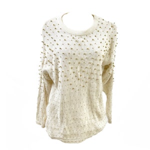 May include: Cream-colored sweater with a crew neck and long sleeves. The sweater is adorned with numerous gold-colored, spherical embellishments scattered across the front and sleeves. The knitwear has a textured appearance.