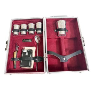 Puede incluir: Maleta plateada abierta con interior de terciopelo rojo, que contiene vasos de chupito plateados, una cuchara, un sacacorchos, naipes y otras herramientas de bar. La maleta está diseñada para guardar accesorios de bar.