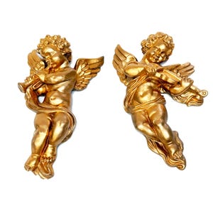 Könnte beinhalten: Zwei goldfarbene Wandskulpturen von Cherubinen. Einer spielt Trompete, der andere Geige. Beide haben detaillierte Flügel und sind mit Stoff drapiert. Die Skulpturen befinden sich auf weißem Hintergrund.
