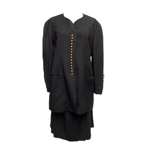 Puede incluir: Un traje de dos piezas negro con una chaqueta larga y una falda. La chaqueta tiene cuello en V, mangas largas y botones dorados en la parte delantera. La falda es hasta la rodilla y plisada. El traje está hecho de tela oscura.