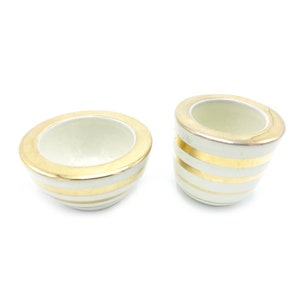 May include: Two ceramic bowls with a cream-colored base and gold horizontal stripes. The rims are also gold. One bowl is round, and the other is cylindrical. The bowls are on a white background.