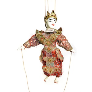 Może przedstawiać: Tradycyjna birmańska marionetka w czerwono-złotym stroju, z koroną i białą twarzą. Marionetka jest zawieszona na sznurkach i gotowa do manipulowania.