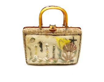 Könnte beinhalten: Vintage rechteckige Handtasche mit gewebtem Strohkörper und transparentem Griff. Die Vorderseite der Tasche ist mit Muscheln, einem Seepferdchen und einem Seestern verziert. Die Tasche hat einen goldfarbenen Verschluss.