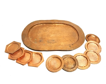 May include: A set of hammered copper serving pieces, including an oval tray and several hexagonal and round coasters. The copper has a warm, reddish-brown hue and a textured surface, suggesting a handcrafted aesthetic. The pieces are arranged on a white background.