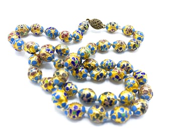 May include: A long necklace made of oval-shaped beads with a blue base and gold accents. The beads are decorated with colorful floral patterns.