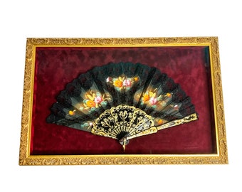 May include: A black lace fan with painted flowers is displayed in a gold-colored frame with a red velvet backing.