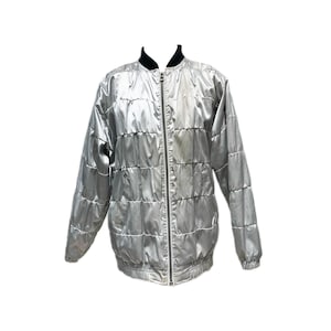 Könnte beinhalten: Silberne Bomberjacke mit Steppdesign. Die Jacke hat einen schwarzen Kragen und einen durchgehenden Reißverschluss. Die Ärmel sind lang und die Jacke scheint aus einem glänzenden, möglicherweise satinartigen Material zu sein.